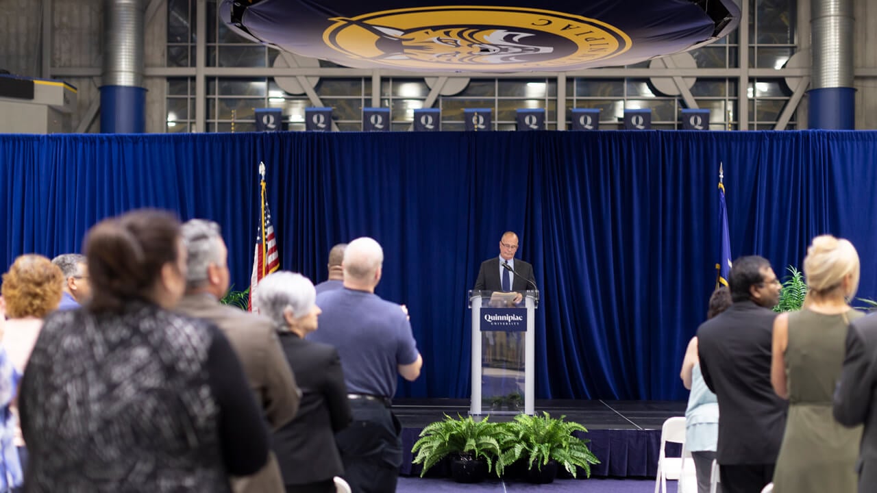 An audience stands and claps while a presenter is at a podium in the basketball arena