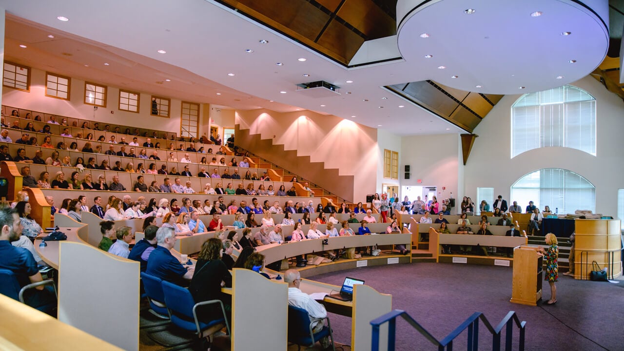 Hundreds of people sit in a large auditorium while a person presents at a podium