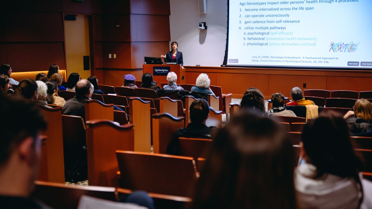 An auditorium filled with people look toward a presenter who is standing in front of a large display