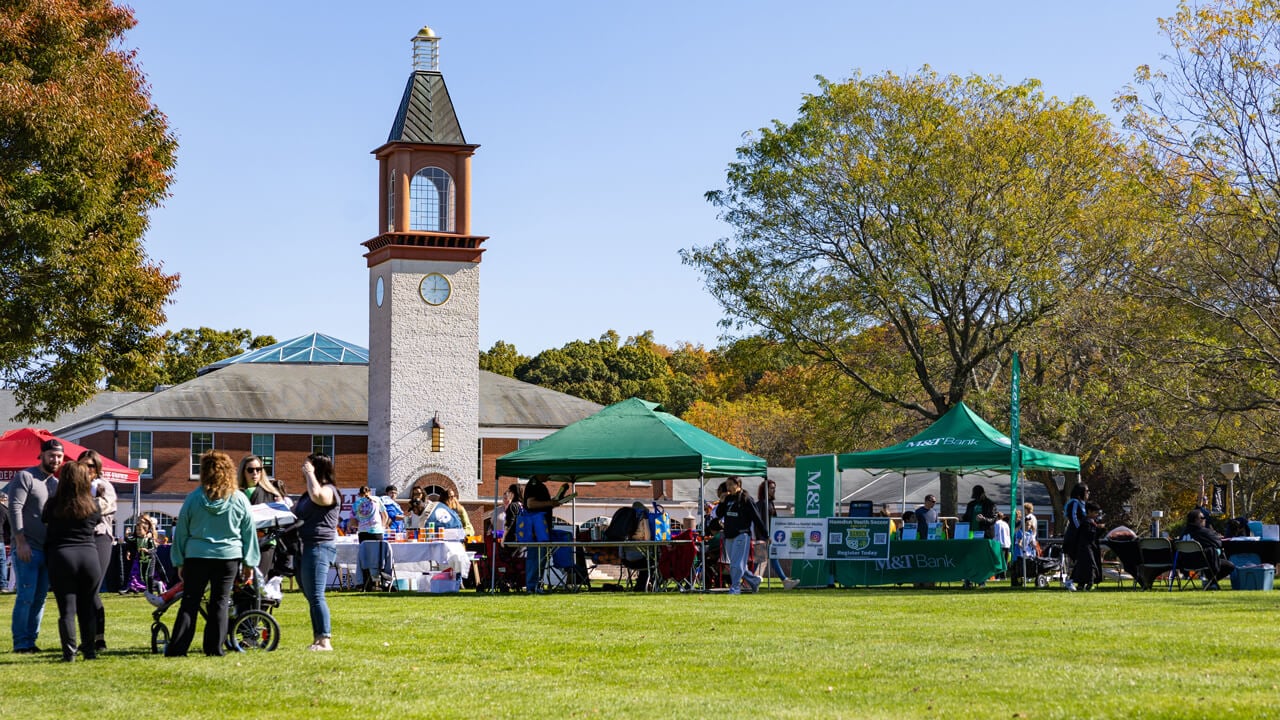 Dozens of people interact on the quad set up with business sun tents with the clocktower in the background