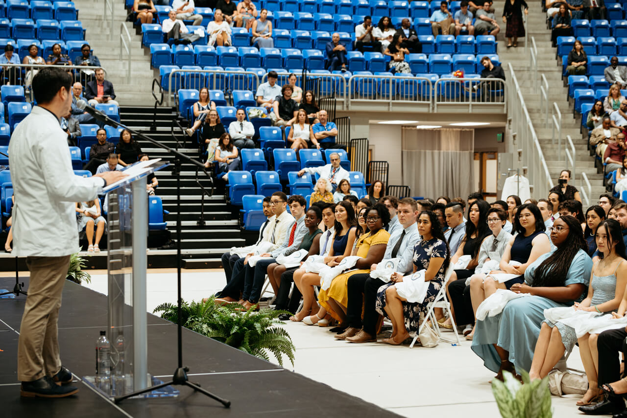 A person presents to rows of audience members on the floor of the arena, with expansive stadium seating behind them